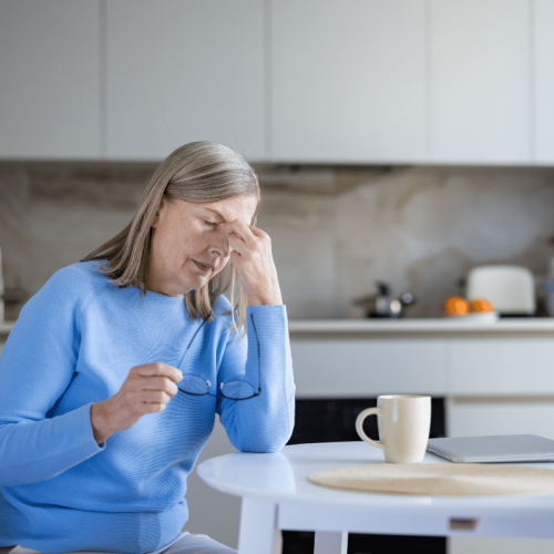 Femme âgée assise à une table de cuisine, se frottant les tempes et tenant des lunettes