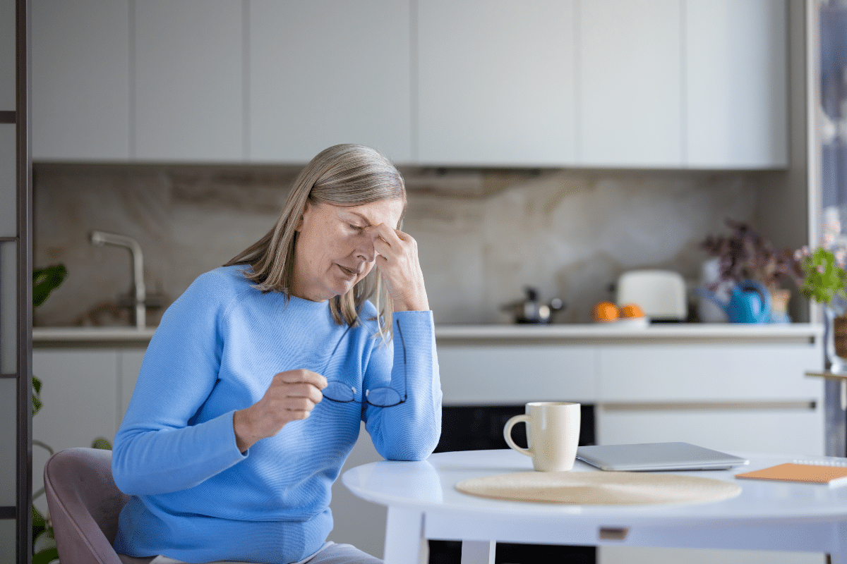 Femme âgée assise à une table de cuisine, se frottant les tempes et tenant des lunettes